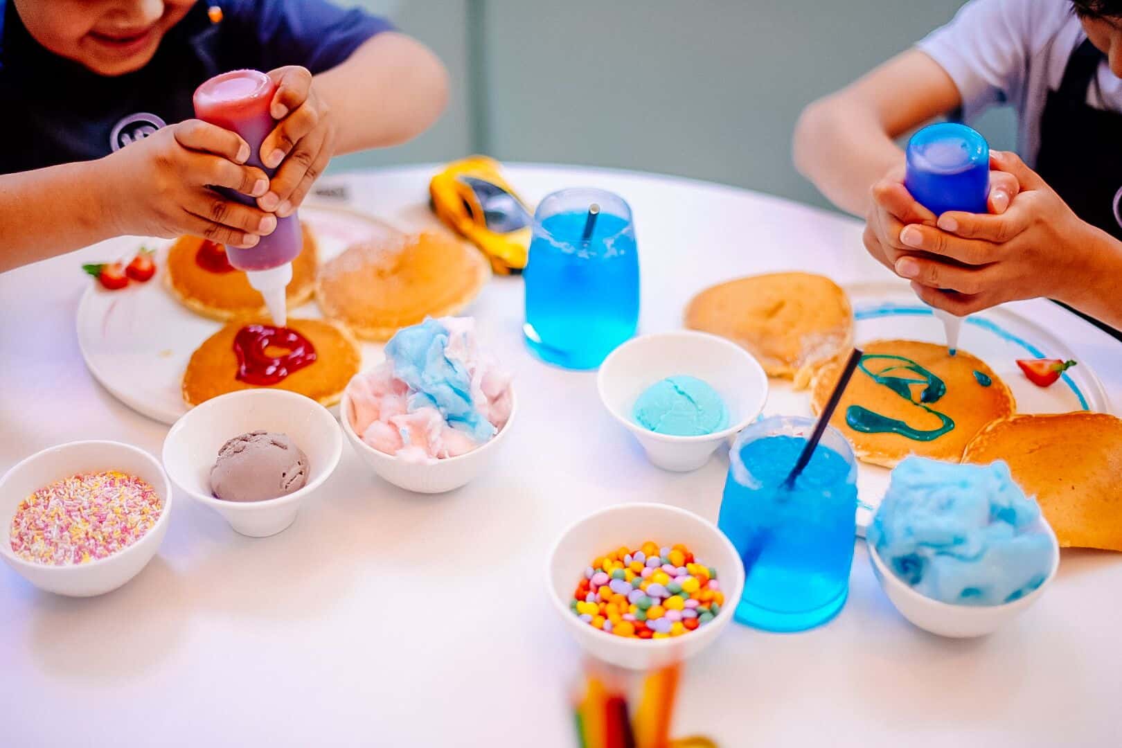 Children decorating pancakes with sauces and sprinkles, with bowls of toppings and blue drinks