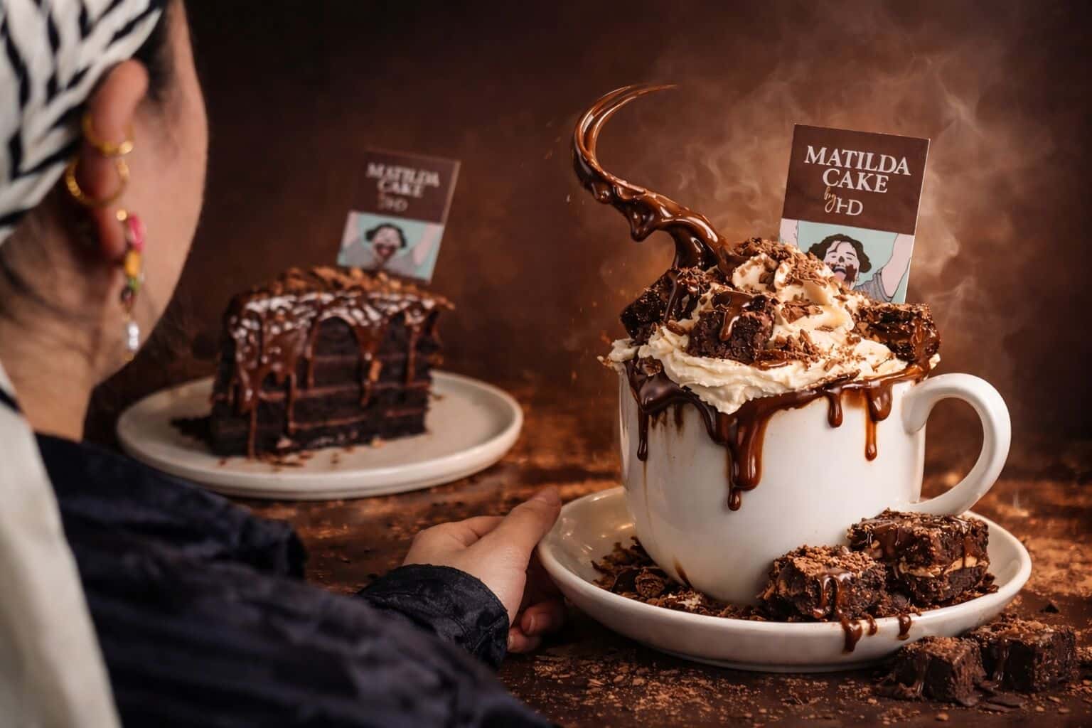 A Matilda hot chocolate piled with cream, chocolate sauce and brownie pieces, beside a slice of Matilda Cake