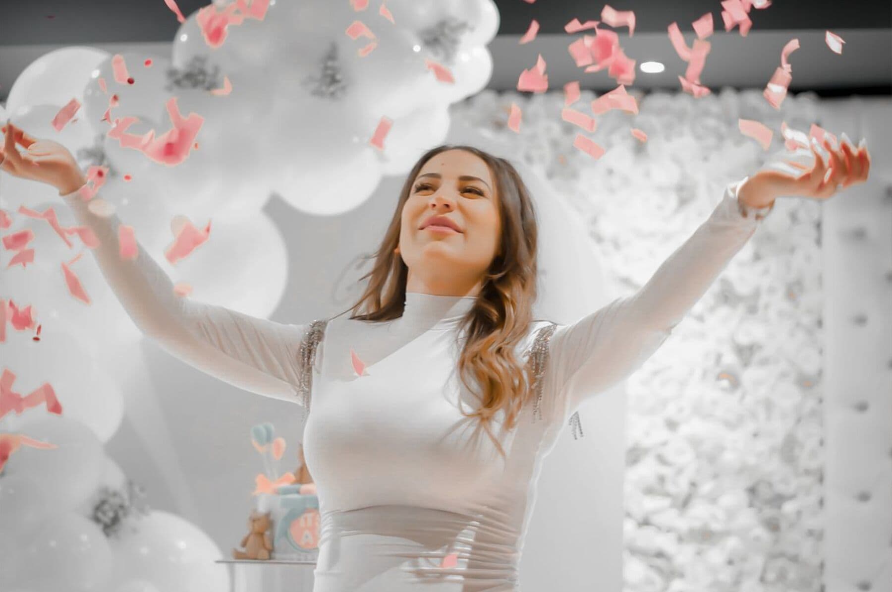 A mother-to-be in cream throwing pink petals in front of a white flower wall