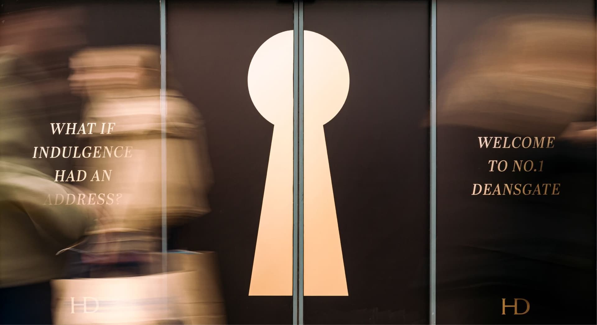 Keyhole-shaped doors glowing gold at the entrance to Haute Dolci Deansgate as shoppers blur past